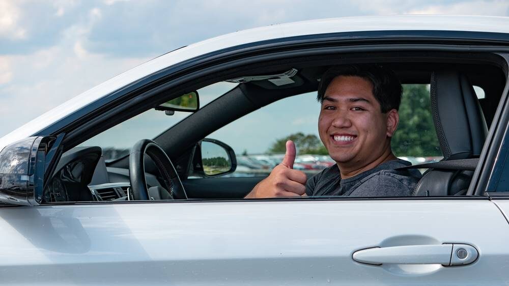 Young student driver sits in drivers seat alone giving a thumbs up.